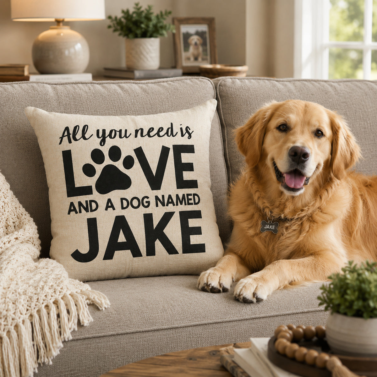 A golden retriever dog lies on a beige sofa next to a cream throw pillow printed with the phrase 'All you need is LOVE AND A DOG NAMED JAKE' in black text and paw-print graphics. The scene is set in a cozy living room with a lamp, books, and small plants in the background.