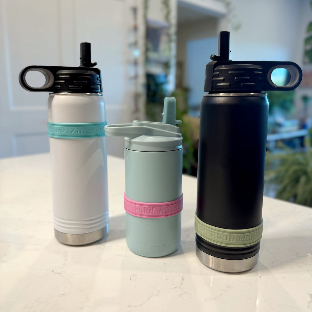 Three water bottles wearing personalized silicone bands: a white bottle with an aqua band, a seaglass blue bottle with a pink band, and a black bottle with a sage band.