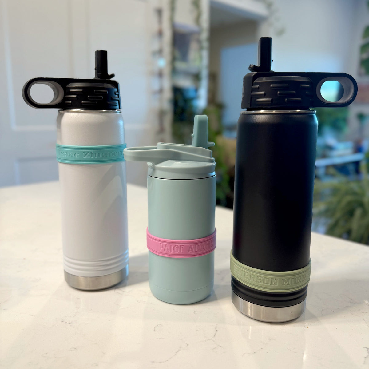 Three water bottles wearing personalized silicone bands: a white bottle with an aqua band, a seaglass blue bottle with a pink band, and a black bottle with a sage band.